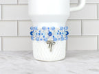 A white tumbler adorned with a beaded bracelet featuring shades of blue and clear beads. A silver bow charm dangles from the bracelet. The tumbler sits on a white and gray surface.
