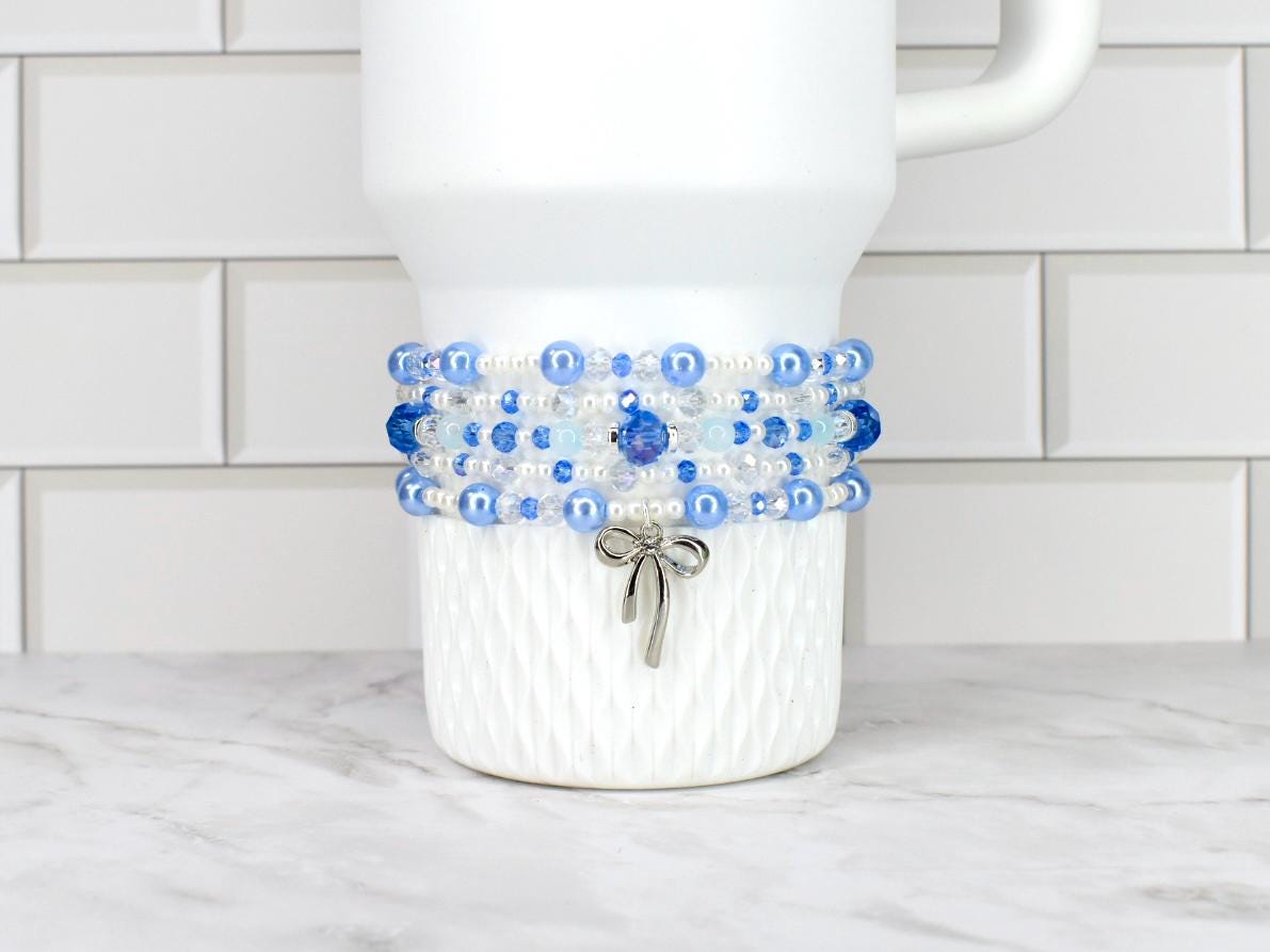 A white tumbler adorned with a beaded bracelet featuring shades of blue and clear beads. A silver bow charm dangles from the bracelet. The tumbler sits on a white and gray surface.