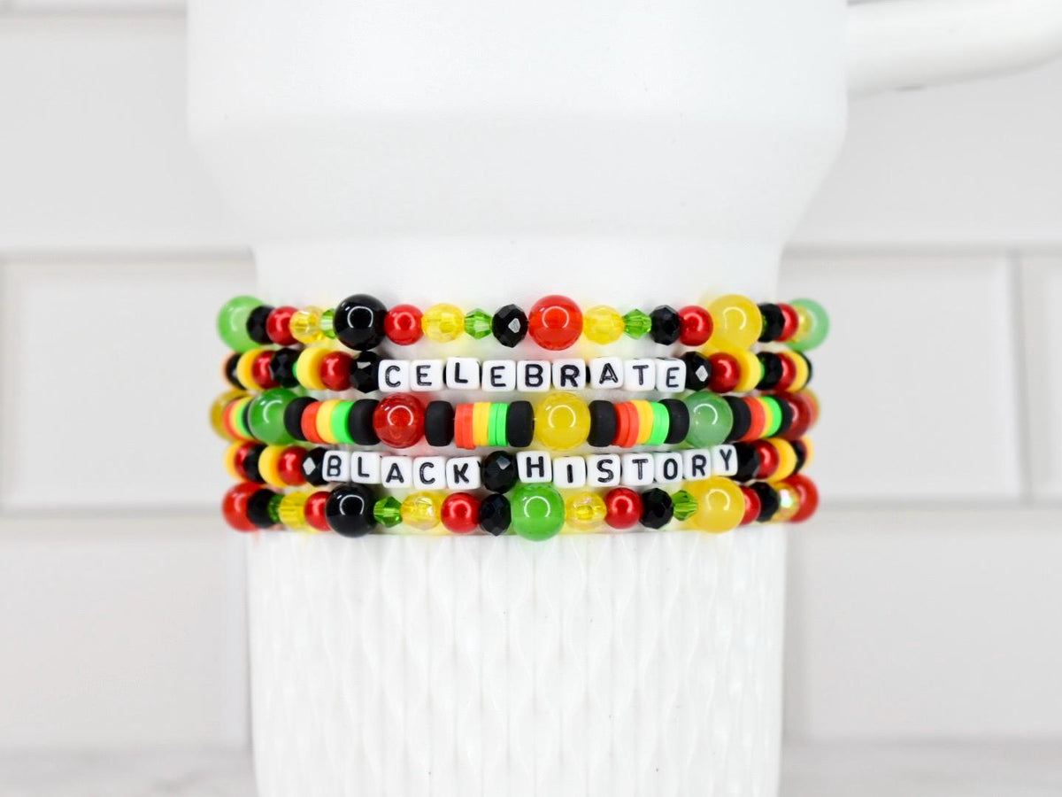 Five beaded bracelets with the words "CELEBRATE BLACK HISTORY" spelled out in white letter beads. The bracelets feature a mix of red, yellow, green, and black beads, and are displayed on a white background.