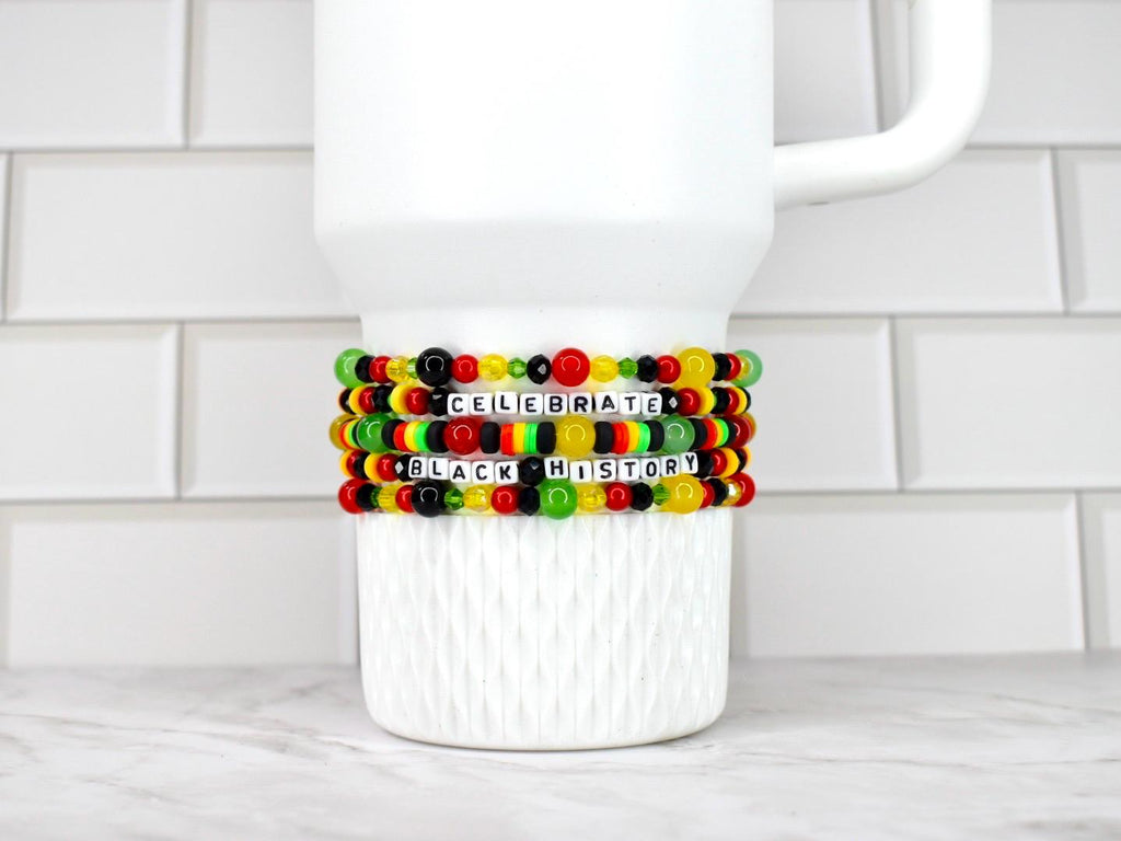 A white Stanley tumbler with a handle, adorned with three beaded bracelets. The bracelets feature black, red, yellow, and green beads, spelling out "CELEBRATE BLACK HISTORY" in white letter beads. The bracelets are stacked on the mug.