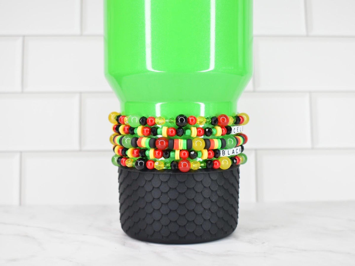 A green tumbler with a black textured base is adorned with several beaded bracelets. The bracelets feature red, yellow, black, and green beads.
