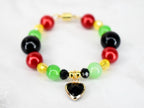 A beaded bracelet featuring red, green, black, and yellow beads. A gold-colored charm with a black heart pendant hangs from the center. The bracelet is secured with a gold-colored clasp.