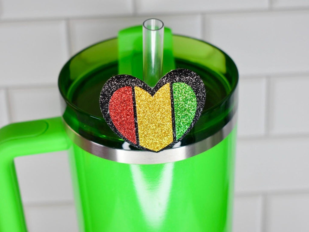 A bright green tumbler with a matching handle and a silver rim. A heart-shaped charm with red, yellow, and green glitter accents sits on the lid. A clear straw is inserted into the lid.