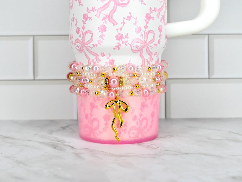 A pink tumbler with a white handle and a decorative beaded wrap. The tumbler has a pink bow pattern and a gold bow charm. The beaded wrap includes pink pearls, clear beads, and gold accents. The tumbler is on a white and gray surface.