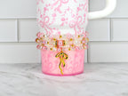 A pink tumbler with a white handle and a decorative beaded wrap. The tumbler has a pink bow pattern and a gold bow charm. The beaded wrap includes pink pearls, clear beads, and gold accents. The tumbler is on a white and gray surface.