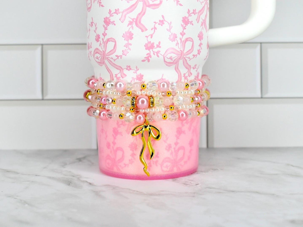 A pink tumbler with a white handle and a decorative beaded wrap. The tumbler has a pink bow pattern and a gold bow charm. The beaded wrap includes pink pearls, clear beads, and gold accents. The tumbler is on a white and gray surface.