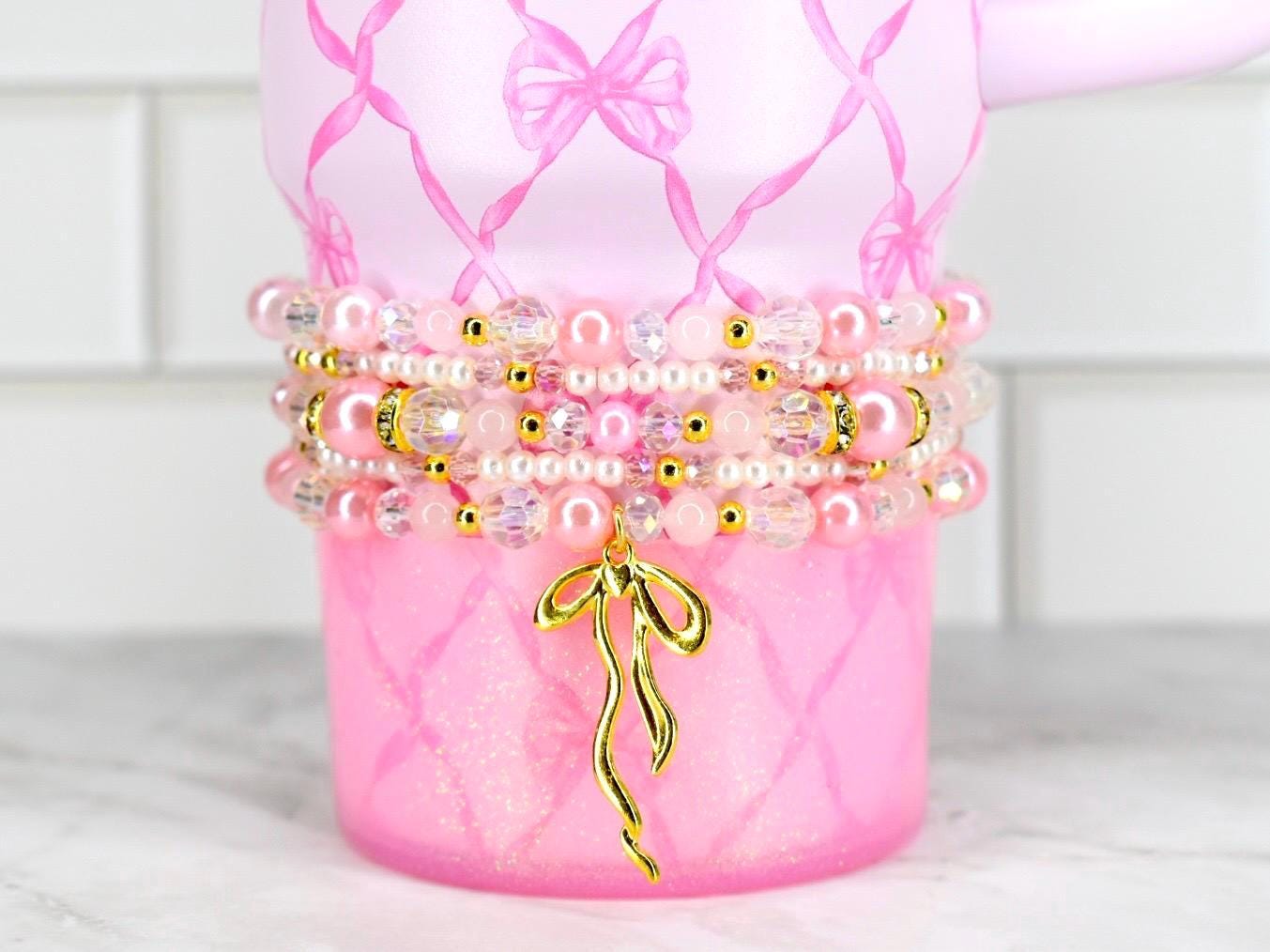Pink tumbler with a bow design and a stack of beaded bracelets. The bracelets feature pink pearls, clear beads, and gold accents. A gold bow charm hangs from the bracelets, adding a touch of elegance.