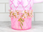 Pink tumbler with a bow design and a stack of beaded bracelets. The bracelets feature pink pearls, clear beads, and gold accents. A gold bow charm hangs from the bracelets, adding a touch of elegance.