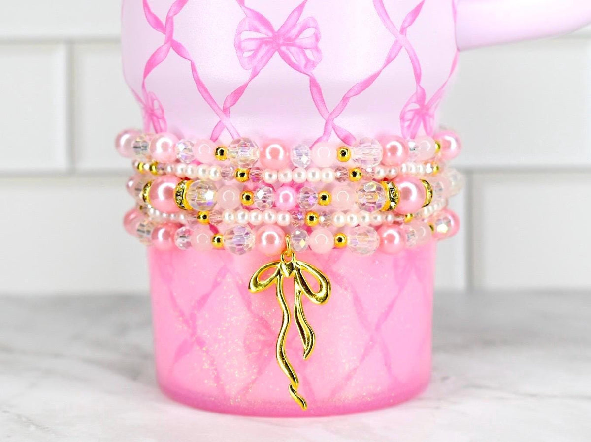 Pink tumbler with a bow design and a stack of beaded bracelets. The bracelets feature pink pearls, clear beads, and gold accents. A gold bow charm hangs from the bracelets, adding a touch of elegance.