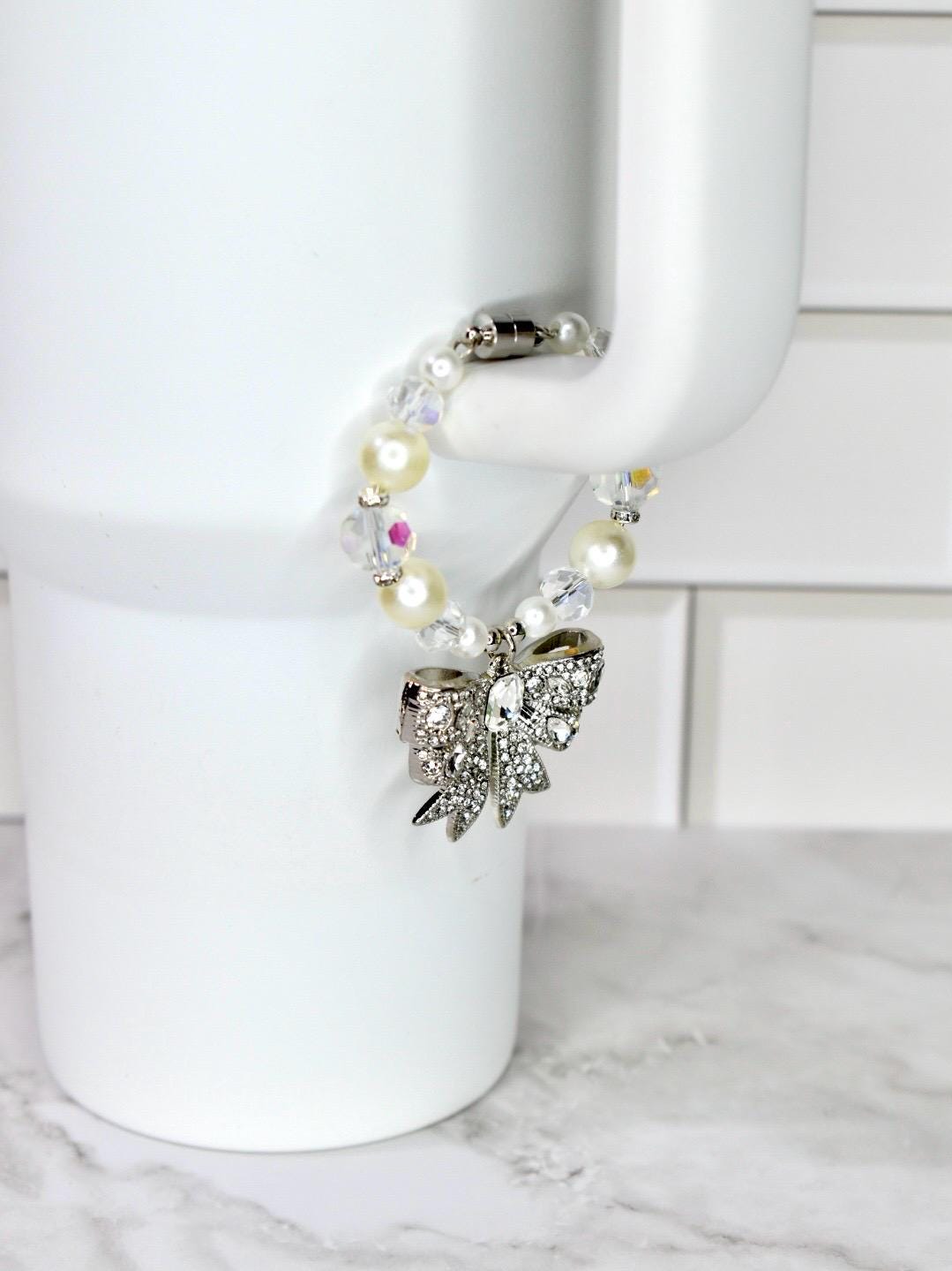 A beaded bracelet with a silver bow charm. The bracelet features a mix of white pearls, clear beads, and iridescent accents. The bow charm is encrusted with clear rhinestones. The bracelet is held by a white mug.