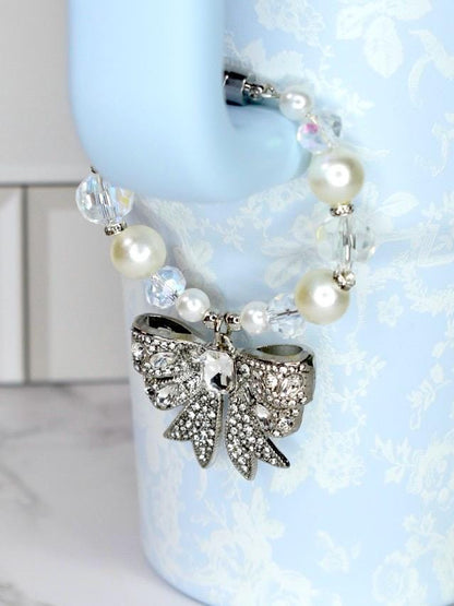 A silver bow-shaped charm with clear rhinestones hangs from a beaded bracelet of white pearls and clear, faceted beads. The charm is attached to a light blue water bottle.