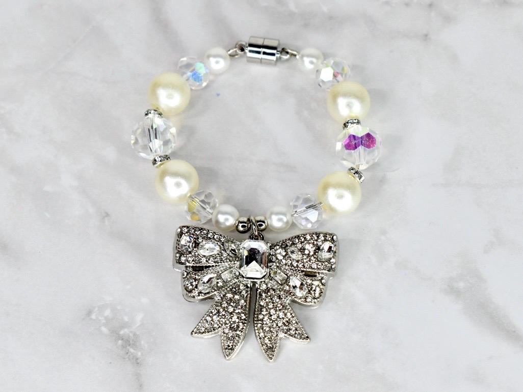 A beaded bracelet featuring cream-colored faux pearls, clear crystal beads, and a silver bow pendant encrusted with rhinestones. The bracelet has a magnetic clasp. A charming accessory.