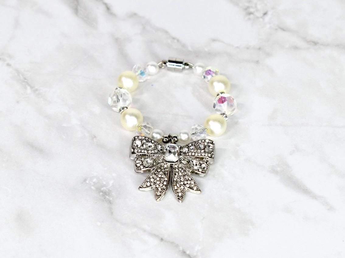 A bracelet featuring a silver bow pendant adorned with clear rhinestones. The bracelet is composed of cream-colored faux pearls and clear, faceted beads. A magnetic clasp secures the bracelet.
