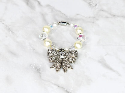A bracelet featuring a silver bow pendant adorned with clear rhinestones. The bracelet is composed of cream-colored faux pearls and clear, faceted beads. A magnetic clasp secures the bracelet.