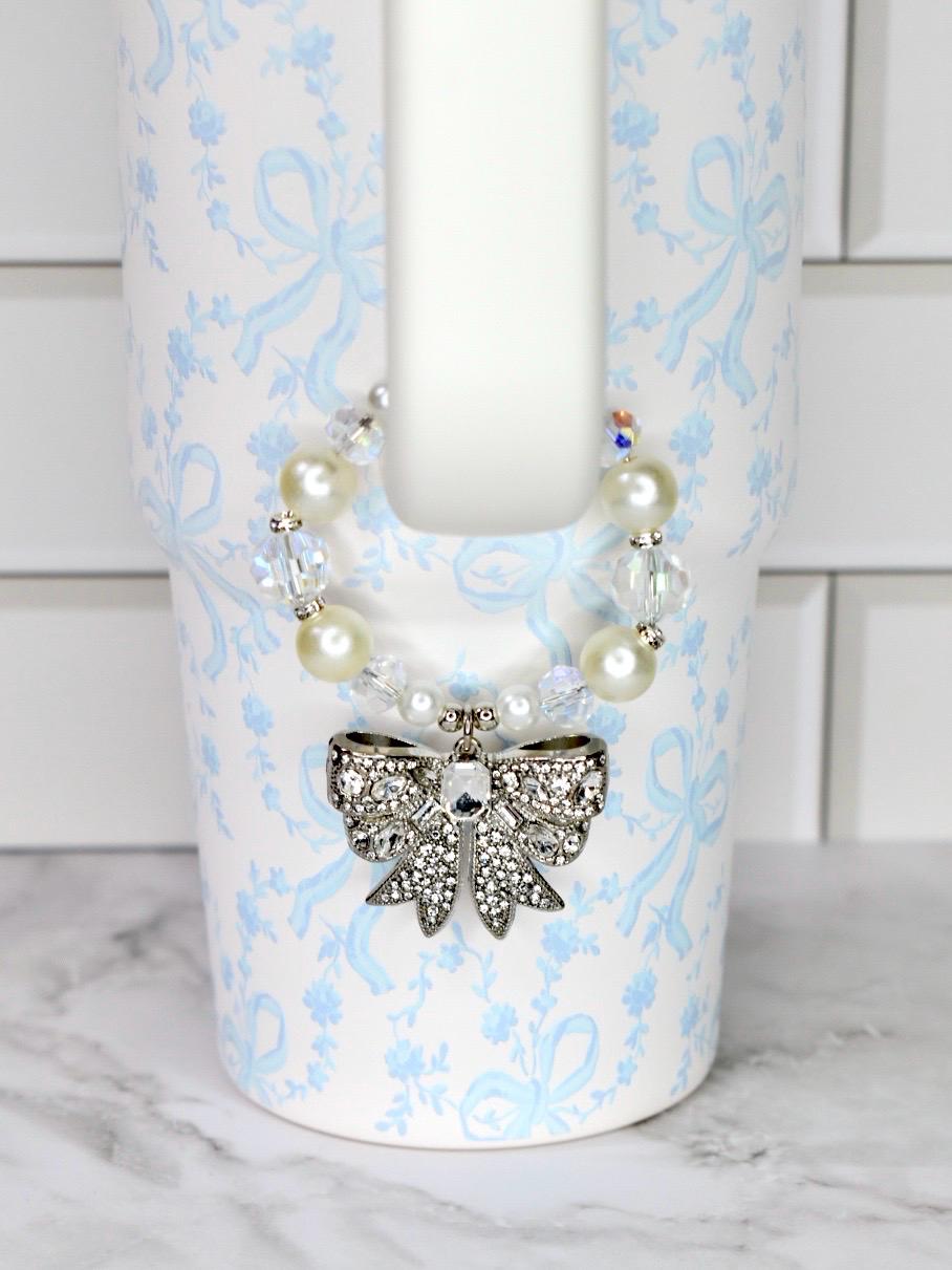 A white LoveShackFancy Stanley tumbler with a light blue floral and bow pattern. A decorative charm with a silver bow pendant and pearl and crystal beads is attached to the tumbler.