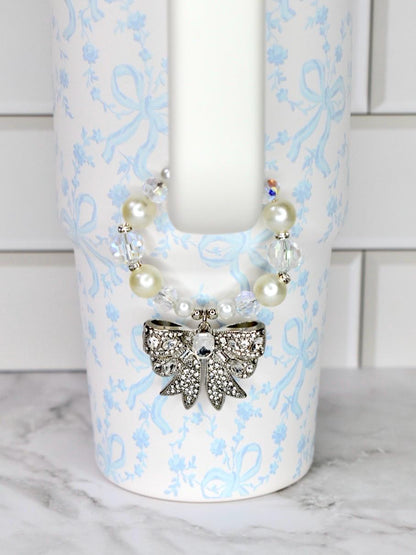 A white LoveShackFancy Stanley tumbler with a light blue floral and bow pattern. A decorative charm with a silver bow pendant and pearl and crystal beads is attached to the tumbler.