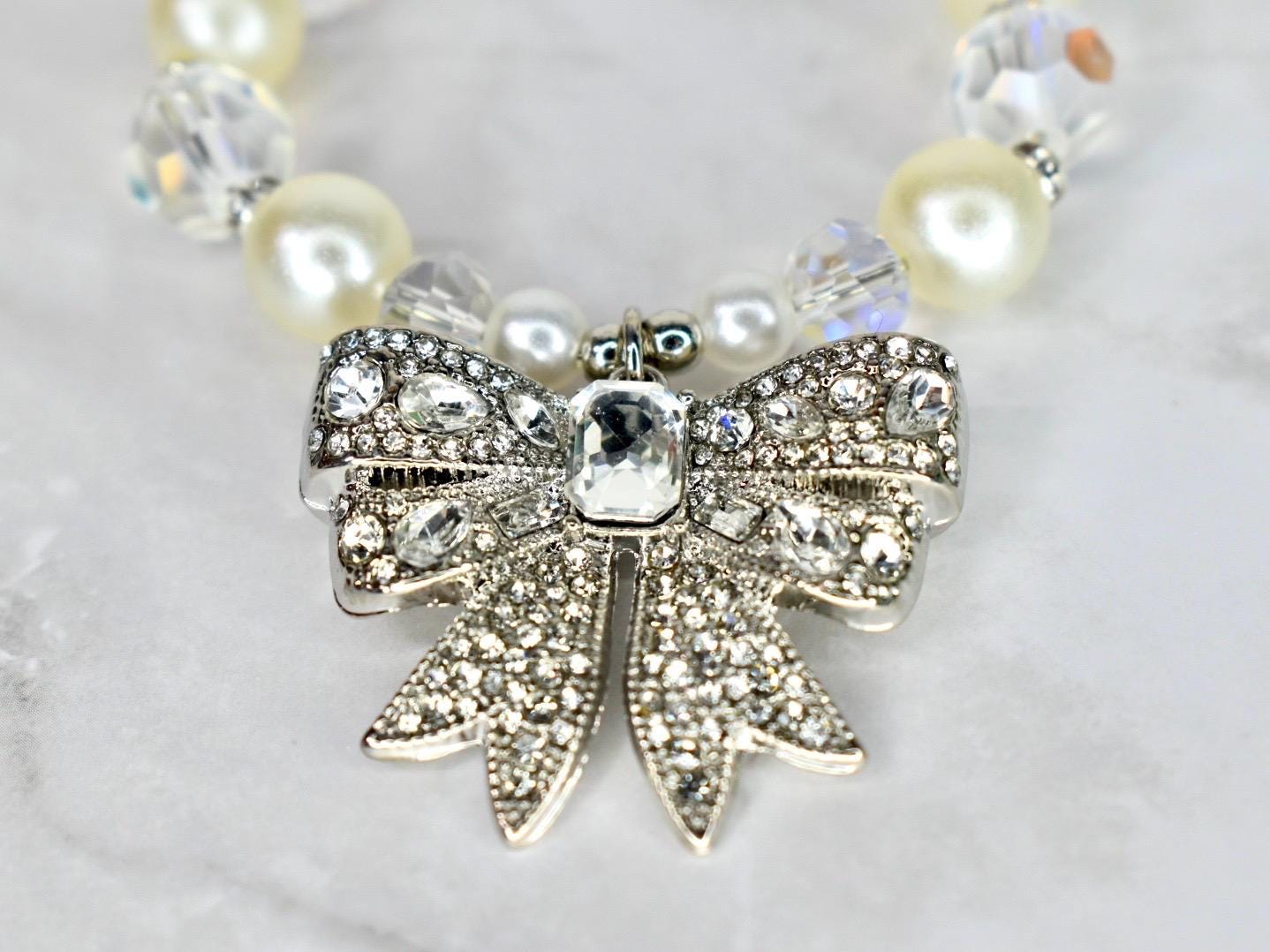 A close-up of a necklace featuring a silver bow pendant encrusted with clear crystals. The bow is the focal point, with a large, rectangular crystal at its center. The necklace includes cream-colored pearls and clear crystal beads.