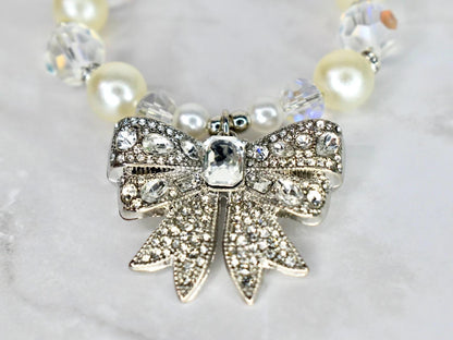 A close-up of a necklace featuring a silver bow pendant encrusted with clear crystals. The bow is the focal point, with a large, rectangular crystal at its center. The necklace includes cream-colored pearls and clear crystal beads.