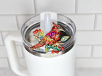 A colorful jeweled cardinal bird straw topper, with red, yellow, and green crystals, is affixed to the lid of a white tumbler. The bird is perched on a branch with red berry accents. The tumbler has a clear lid and a white handle.