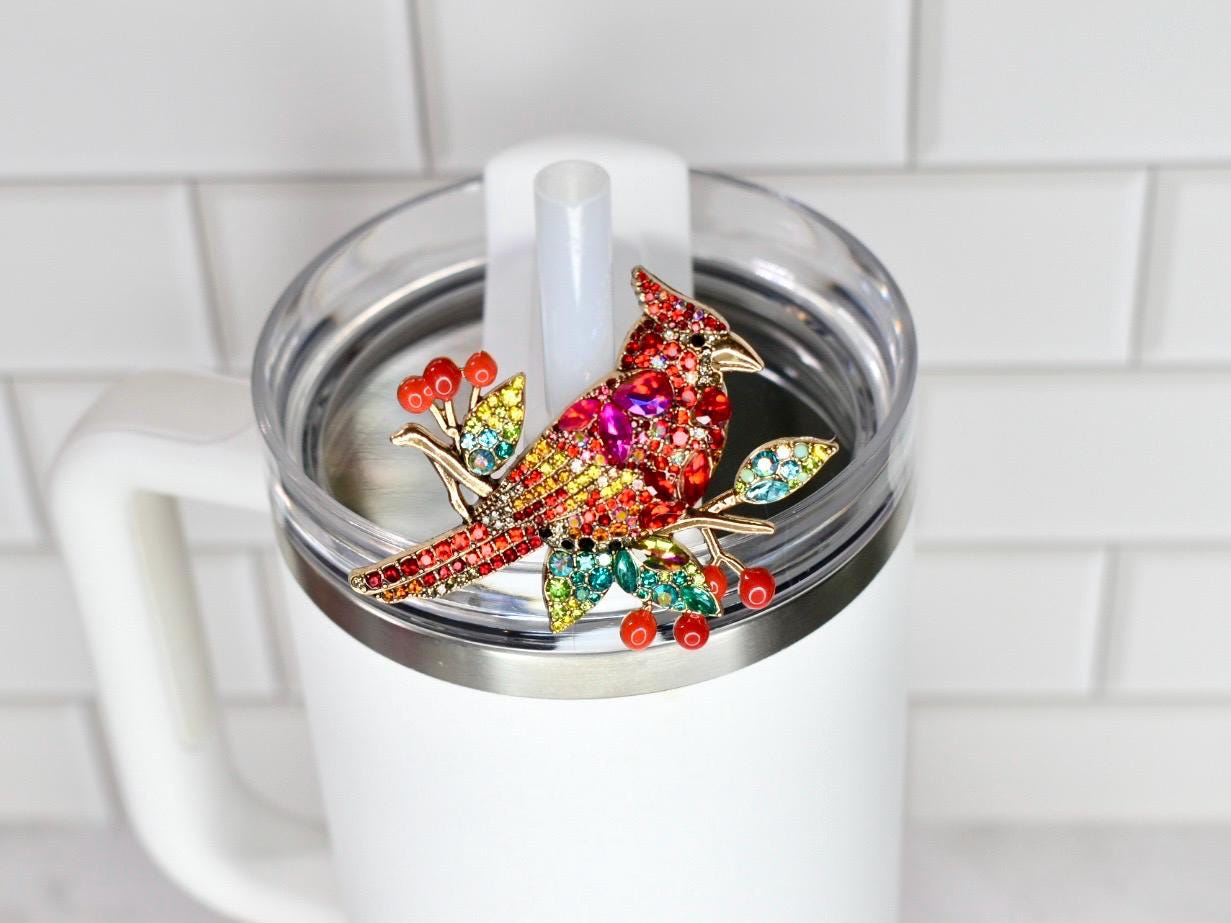 A colorful jeweled cardinal bird straw topper, with red, yellow, and green crystals, is affixed to the lid of a white tumbler. The bird is perched on a branch with red berry accents. The tumbler has a clear lid and a white handle.