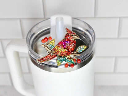 A colorful jeweled cardinal bird straw topper, with red, yellow, and green crystals, is affixed to the lid of a white tumbler. The bird is perched on a branch with red berry accents. The tumbler has a clear lid and a white handle.