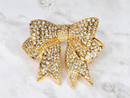 A gold-toned bow brooch encrusted with clear crystals. The bow design features a central knot and two symmetrical loops with pointed ends. The brooch is set against a marble background.