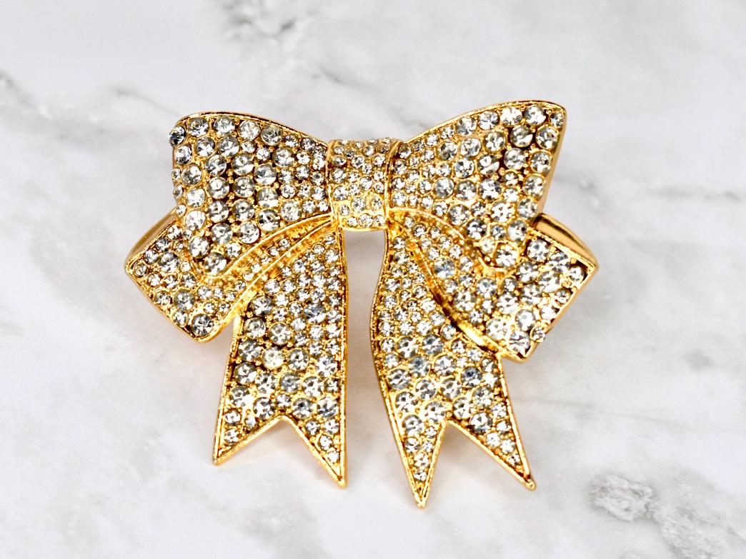 A gold-toned bow brooch encrusted with clear crystals. The bow design features a central knot and two symmetrical loops with pointed ends. The brooch is set against a marble background.