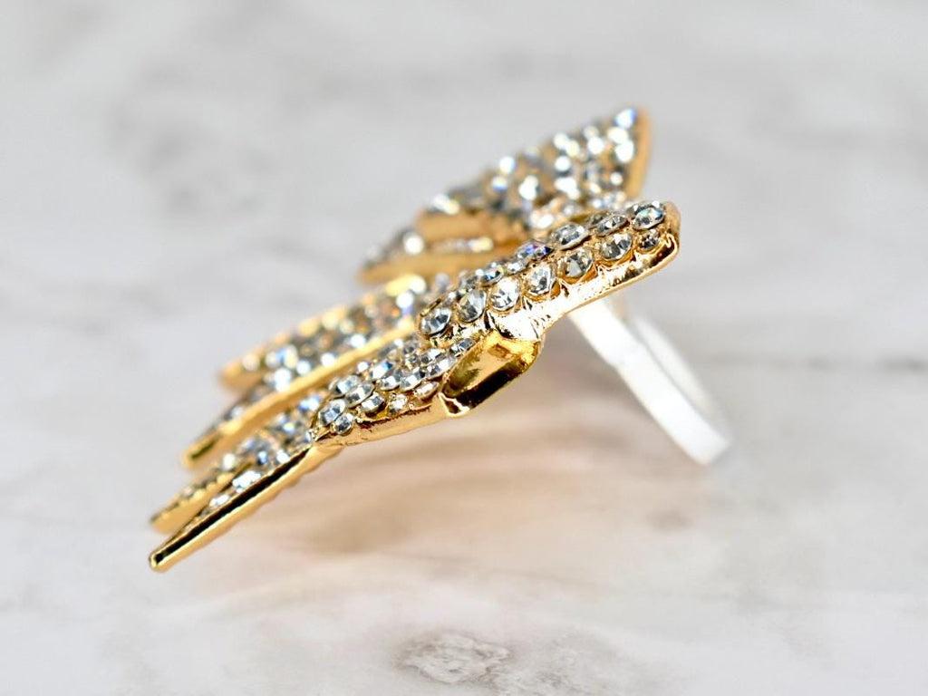 A gold-colored ring with a wing-like design, encrusted with numerous small, clear crystals. The ring's band is a simple, polished silver. The wings are angled, creating a dynamic and eye-catching appearance. The ring is set against a soft, marbled background.