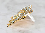 A gold-colored ring with a wing-like design, encrusted with numerous small, clear crystals. The ring's band is a simple, polished silver. The wings are angled, creating a dynamic and eye-catching appearance. The ring is set against a soft, marbled background.