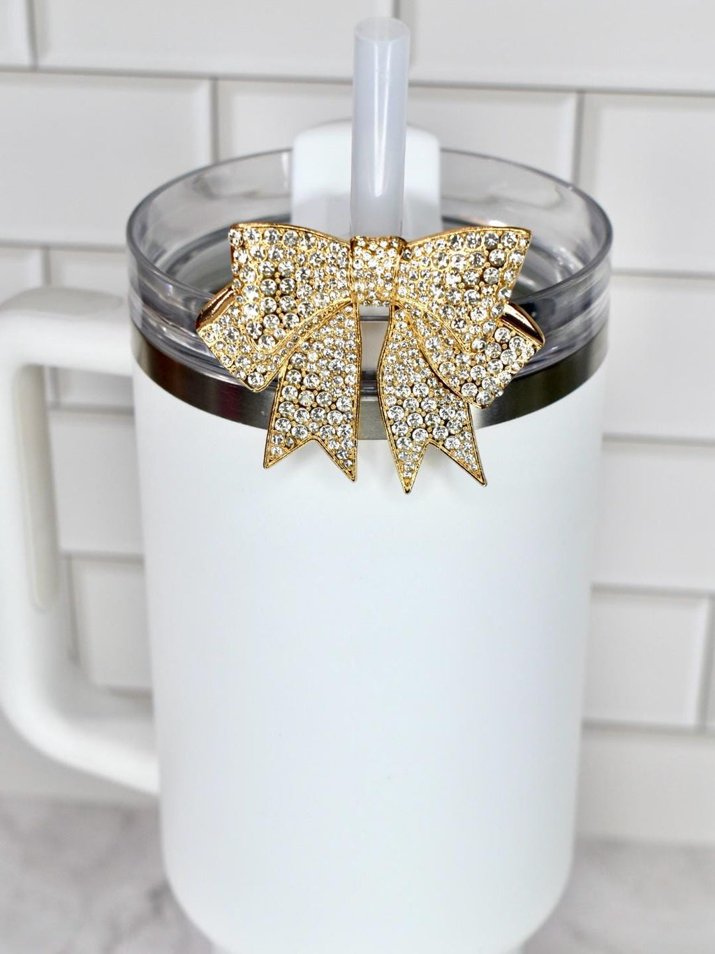 A white tumbler with a clear lid and a gold bow-shaped charm adorned with sparkling crystals. A white straw is inserted through the lid. The tumbler has a white handle.