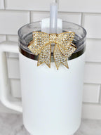 A white tumbler with a clear lid and a gold bow-shaped charm adorned with sparkling crystals. A white straw is inserted through the lid. The tumbler has a white handle.