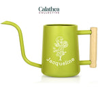 A bright green metal watering can with long gooseneck spout and wood handle with a white flower bouquet design and 'Jacqueline' below the bouquet in white permanent vinyl.