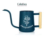 A navy blue metal watering can with long gooseneck spout and wood handle with a white flower bouquet design and 'Jacqueline' below the bouquet in white permanent vinyl.