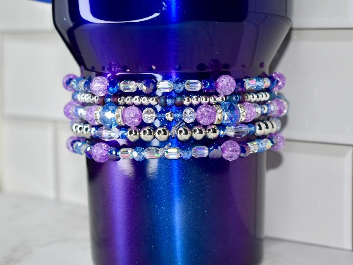 A deep blue tumbler with a gradient finish, adorned with multiple rows of beaded embellishments. The beads are a mix of purple, silver, and blue, creating a sparkling, decorative effect. The tumbler is designed for beverages.