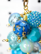 A cluster of blue and turquoise beads with gold-colored accents. The beads vary in size and texture, including faceted, glittery, and marbled designs. The beads are connected with gold-colored jump rings and chains.