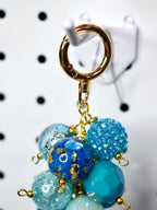 A gold-colored keychain with a cluster of blue and turquoise beads. The beads vary in texture, including faceted, glittery, and marbled designs. Some beads have gold leaf accents. The keychain is attached to a gold clasp.