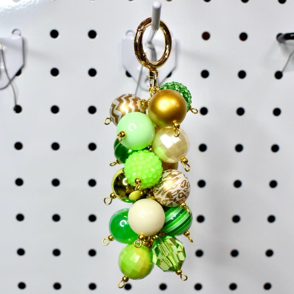A beaded keychain with a gold clasp. The keychain features a cluster of various green, gold, and cream-colored beads. Some beads are solid, while others have patterns or textures. The beads are connected with small gold-colored loops.