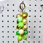 A beaded keychain with a gold clasp. The keychain features a cluster of various green, gold, and cream-colored beads. Some beads are solid, while others have patterns or textures. The beads are connected with small gold-colored loops.