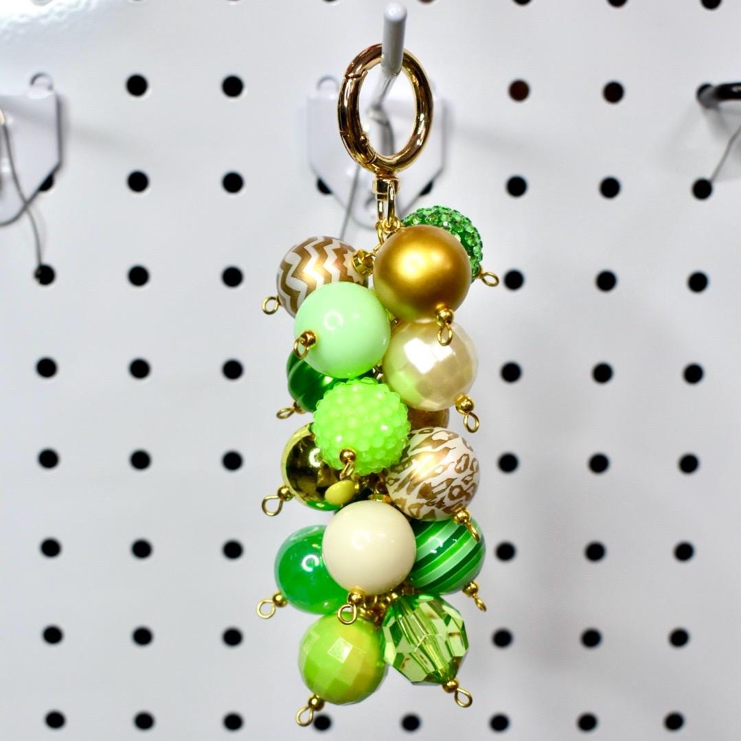A beaded keychain with a gold clasp. The keychain features a cluster of various green, gold, and cream-colored beads. Some beads are solid, while others have patterns or textures. The beads are connected with small gold-colored loops.