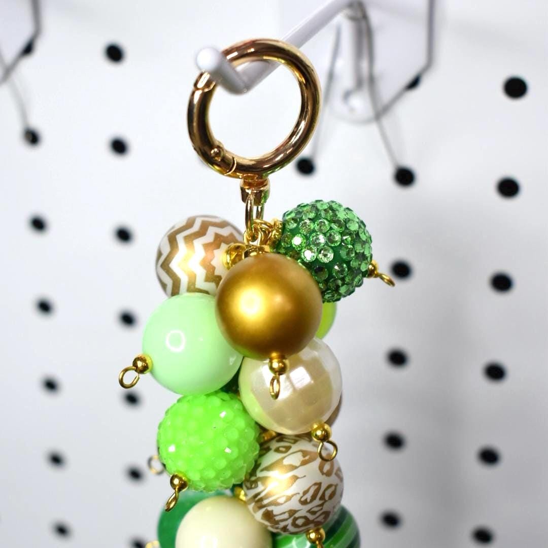 A beaded keychain with a gold clasp and a cluster of colorful beads. The beads are in shades of green, gold, and cream, with some featuring textured and patterned designs. The keychain is hanging against a white background.