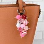 A pink and red beaded charm hangs from a brown handbag. The charm features various sizes of pink, white, and red beads, including striped and crystal-like designs. The handbag has a smooth, leather-like texture.