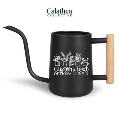 Black metal watering can with a long, curved spout and a wooden handle. The can features a white graphic of potted plants and the text "Custom Text" and "Optional Line 2". The logo "Calathea Collective" is at the top.
