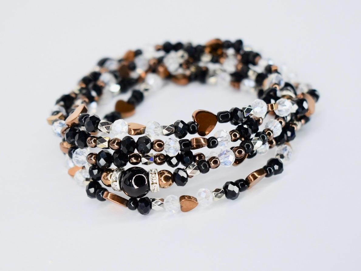 A multi-strand wrap bracelet featuring a mix of black, clear, and bronze-colored beads. The bracelet includes heart-shaped and round beads, with a central black bead accented with silver-colored details. The bracelet is designed to wrap around the wrist multiple times.
