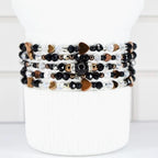A stack of beaded bracelets in black, white, and gold tones. The bracelets feature heart-shaped charms and various bead sizes. The bracelets are displayed on a white cylindrical stand.