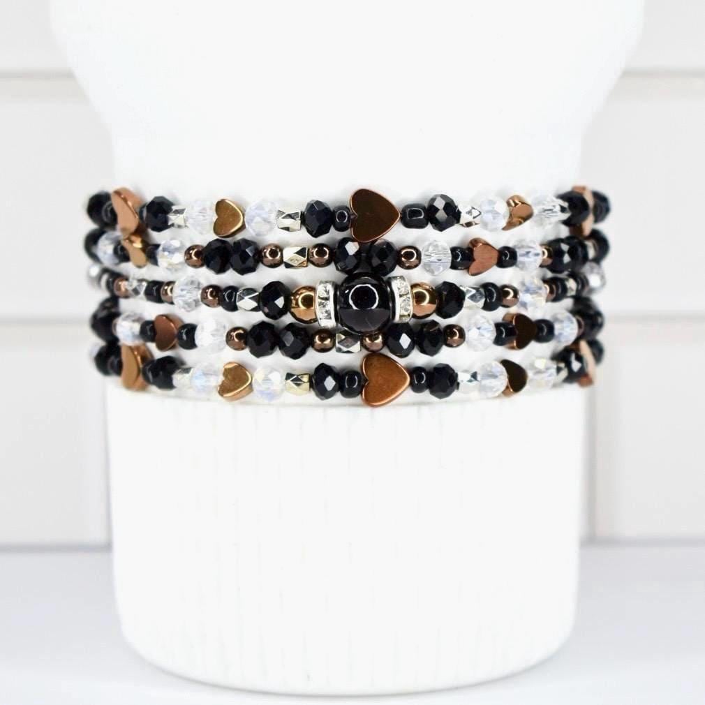 A stack of beaded bracelets in black, white, and gold tones. The bracelets feature heart-shaped charms and various bead sizes. The bracelets are displayed on a white cylindrical stand.