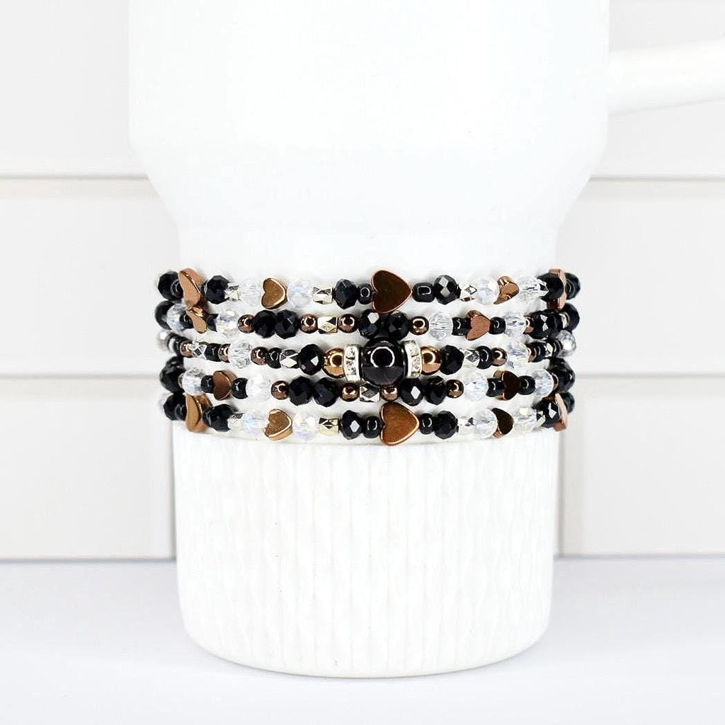 A stack of beaded bracelets in black, white, and gold tones. The bracelets feature heart-shaped and round beads. The bracelets are wrapped around a white cylindrical object.