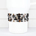 A stack of beaded bracelets in black, white, and gold tones. The bracelets feature heart-shaped and round beads. The bracelets are wrapped around a white cylindrical object.