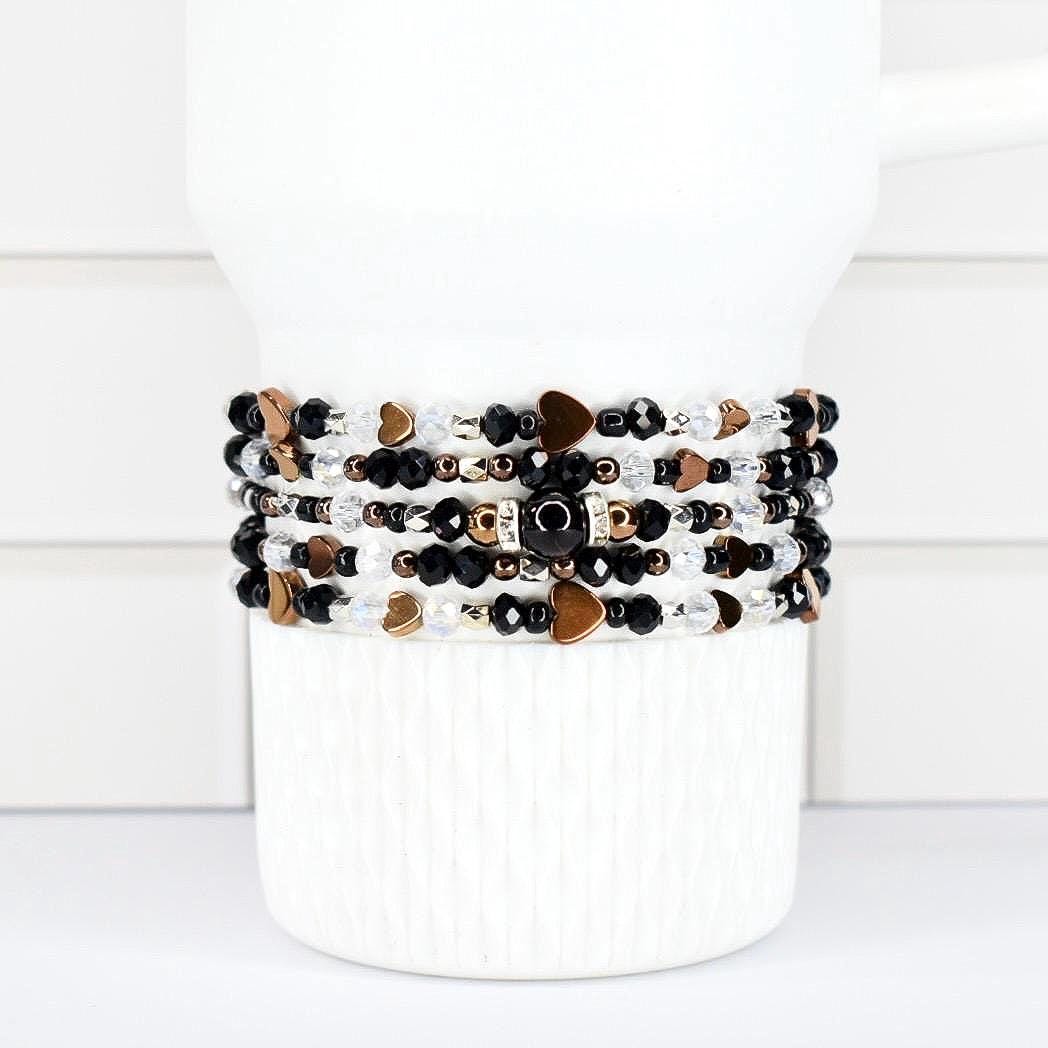 A stack of beaded bracelets in black, white, and gold tones. The bracelets feature heart-shaped and round beads. The bracelets are wrapped around a white cylindrical object.
