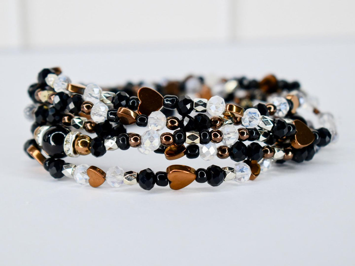 A multi-strand bracelet with black, clear, and bronze beads. The bracelet features heart-shaped charms and metallic accents. The beads are strung together to create a wrap-around design, suitable for wrist sizes of various sizes.