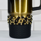 A gold-colored travel mug with a black handle. The mug is adorned with multiple layers of black and gold beaded accents. The bottom of the mug features a textured black rubber sleeve.
